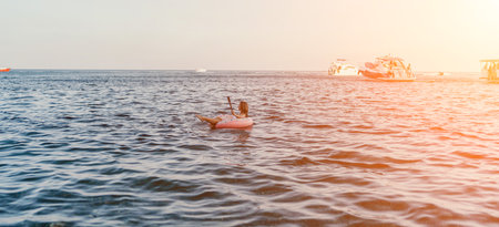 Woman, floating, ocean. Young woman relaxes on inflatable donut in open sea at golden hour with boats.の写真素材