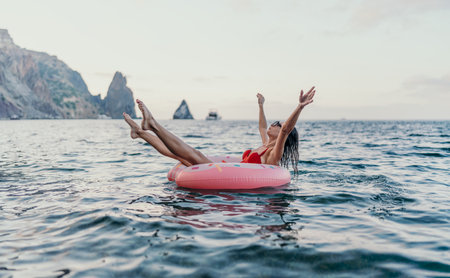Woman, floating, sea. Happy woman on a pink donut ring in the open sea, enjoying summer vacation by majestic cliffs.の写真素材