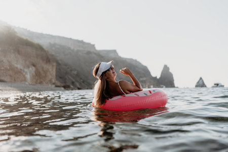Woman, ocean, donut. Young woman floats on a pink donut ring enjoying summer relaxation by rocky cliffs, copy spaceの写真素材