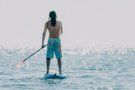 Man Paddleboarding Ocean Water Sport - A man stands on a paddleboard on the water, paddling with an...