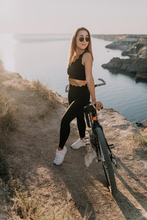 Woman bicycle cliff standing on a high ridge overlooking the ocean at sunset with a mountain bike representing fitness and outdoor adventureの写真素材