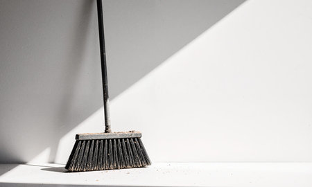 Sweeping Tool Stillness. A wide, industrial push broom leans against a flawless white cyc wall, its bristles frayed and dustedの素材