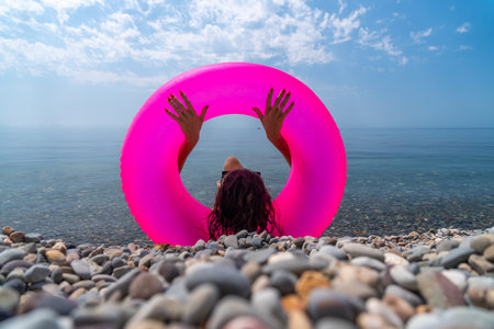 Beach vacation inflatable woman sits on a pebble beach holding a bright pink swim ring facing the...