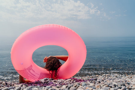 Inflatable ring beach Woman sits on a pebble shore behind a bright pink floaty looking at the calm blue seaの写真素材