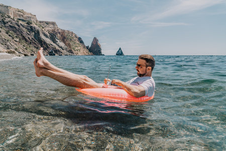 Freelancer laptop ocean man works on a computer while floating on an inflatable ring in the sea near rocky coastal cliffs during summerの写真素材