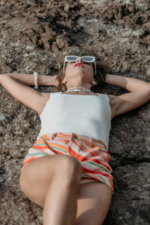 Woman relaxation summer young woman wearing white sunglasses and striped shorts lying on a rocky surface during a sunny dayの写真素材