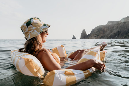 Woman Inflatable Raft Sea Relaxation - A woman wearing a hat and sunglasses relaxes on an inflatable raft in the sea, with rocky cliffs in the background.の写真素材