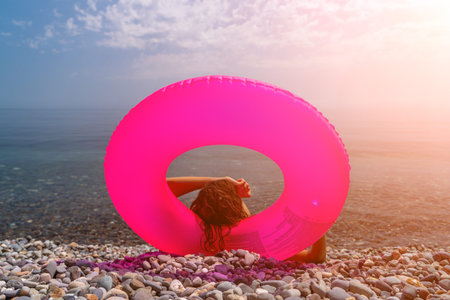 Inflatable beach vacation woman relaxes on a pebble shore framed by a bright pink donut float looking toward the calm blue seaの写真素材