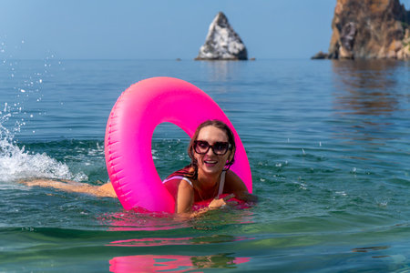 Woman swimming ocean pink inflatable ring smiling in turquoise water with rocky cliffs background...
