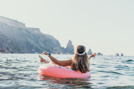 Woman floating ocean, relaxing on pink donut ring in clear water with scenic cliffs and copy space.