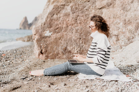Laptop beach work woman sitting on rocky shore typing on computer wearing striped sweater for remote digital nomadの写真素材