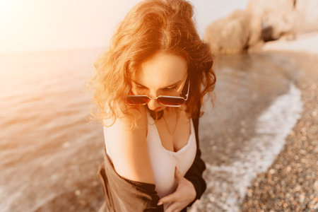 Woman sunset beach wearing sunglasses and a white top stands on a rocky shore during a golden hour...