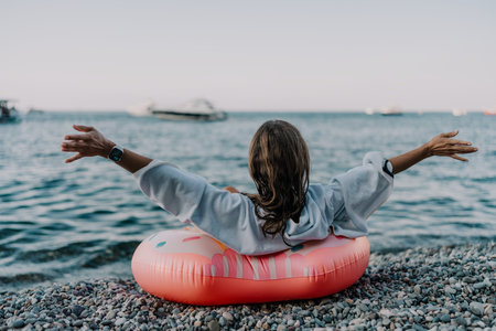 Woman, beach, ocean. Young woman on pebble beach with inflatable ring, arms outstretched, enjoying...