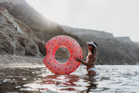 Woman, beach, donut. Happy woman in white bikini holding donut float at rocky beach during sunset.