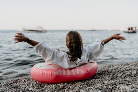 Woman, beach, ocean. Relaxed woman on donut float stretches arms on pebble beach looking at sea.