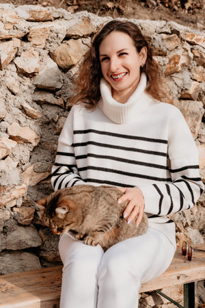 Woman cat sweater smiling while sitting on bench outdoors with her pet tabby cat against rustic...