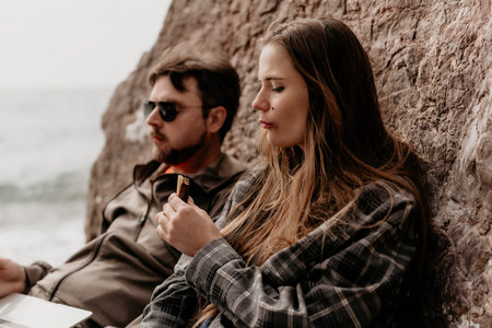 Couple coastal relaxing resting near rocks while woman eats a snack and man wears sunglasses on a...