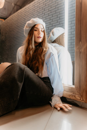 Woman mirror beret sitting on a floor in front of a mirror wearing a blue shirt and dark jeans in a...