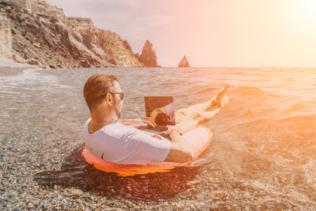 Freelancer working laptop man floating on inflatable ring in ocean water by rocky cliffs during...