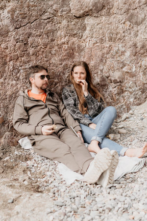 Beach couple picnic sitting against a steep rocky cliff enjoying a snack outdoors during a relaxing...