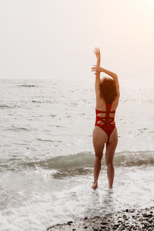 Woman beach swimsuit enjoying the ocean waves at sunset with arms raised in a gesture of freedom and serenityの写真素材