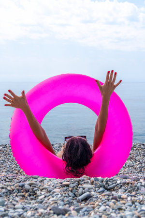 Woman floating relax, pink inflatable ring, raising arms to the summer sky at a pebble beach.