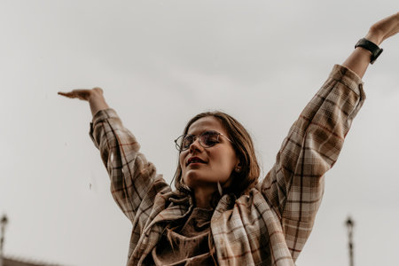 Woman rain freedom. Joyful young woman with glasses standing in the rain with arms raised towards the gray sky