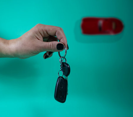 Female Hand with Car Keys on a Turquoise Background. Close-up Of A Hand Holding A Car Key With A Red Car In The Background. The Seller Hands over the Keys. Car Buying Concept.の写真素材