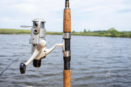 Close-up of a Fishing rod Wheel Against the Backdrop of a Fantastic Lake with Perch, Carp and Pike. Fishing on the Lake in the Morning at Sunrise or in the Evening at Sunset. fishing background.の写真素材