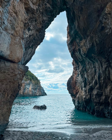 Natural rock arch frames a peaceful scene of the ocean and coastline. Waves gently lap at the shore while clouds drift in the sky. This coastal view is serene and inviting. San Nicola Arcella, Calabria, Italy.の写真素材