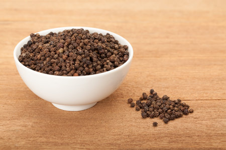 black peppercorns in white ceramic bowl on wood background