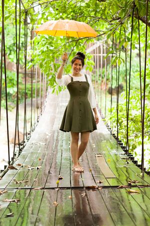 Portrait of beautiful asian woman is standing on wooden suspension bridge with yellow umbrella in rainy dayの写真素材