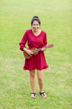 Portrait of beautiful young asian woman with red dress is playing ukulee and feeling happy and smile on green field at the public parkの写真素材