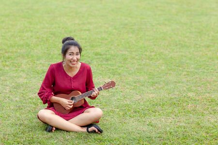 Portrait of beautiful young asian woman with red dress is playing ukulee with feeling happy and smile on green field at the public parkの写真素材