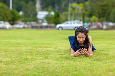 Portrait of beautiful young asian woman lie on your stomach and using smart phone and smile on green grass field at the public parkの写真素材