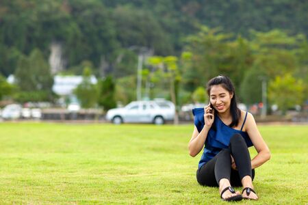 Portrait of a happy asian woman talking on mobile phone while sitting on green grass field in the public parkの写真素材