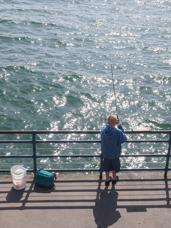 CALIFORNIA, UNITED STATES - April 1, 2012 : Boy in blue shirt stand on a pier with a fishing rod by the seaのeditorial素材
