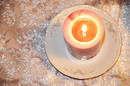A burning white pink candle on a porcelain saucer, standing on a silver cloth.の写真素材