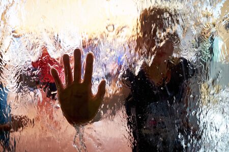 A hand pressed to the glass, with a stream of waterfall on the surface of the glass. Water flows through the glass, Glass closeup. Abstract background blurred by water flows.の写真素材
