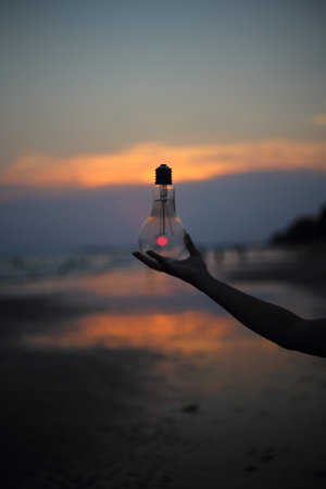 hand holding a electric light bulb and sun at beach sunset sky
