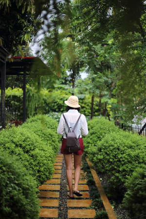 Back view of young Asian woman wearing hat and backpack walking in garden.の写真素材