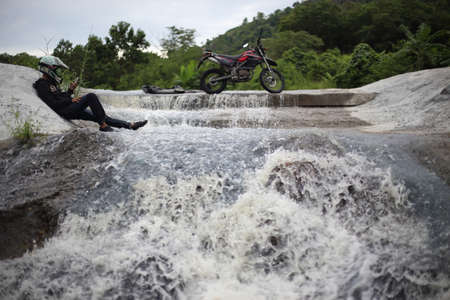 Rear view of a man sitting on a rock next to a river with a motorbike parked nearby.の写真素材