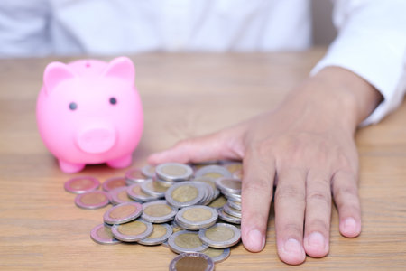 Hand putting coin to Pink Piggy bank and Thai coins stacks on the wooden table. Save money for...
