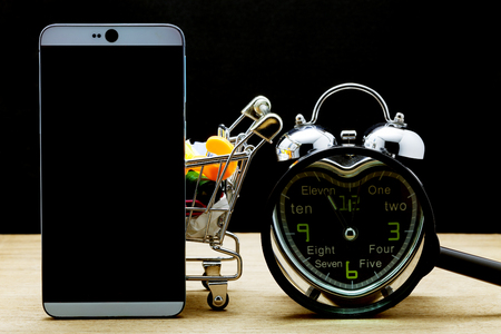 Smartphone with alarm clock and shopping cart on wooden floor,time to online shopping concept の写真素材