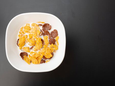 Breakfast is ready to be eaten in a white bowl consisting of cornflakes, crispy chocolate, and milk, placed on a black painted metal dining table.の写真素材