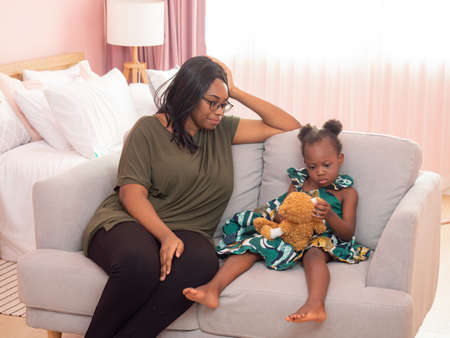 Head shot portrait of happy African American mother cuddles a lovely little daughter sitting on the sofa and looking at camera.の写真素材
