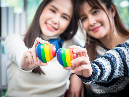 Selective focus, young Asian lesbian couple holding a rainbow heart symbolizes the LGBT community, in the living room at home, the lifestyle of couple LGBT loving women happy in romantic love and funの写真素材