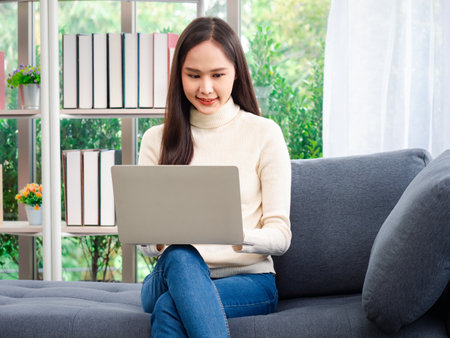 Attractive young beautiful Asian women stay at home with a laptop doing research work for her business while sitting on the sofa in the living room.の写真素材