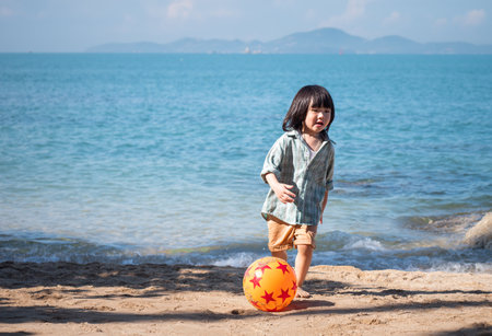 Happy Asian little boy playing barefoot soccer on the beach with a red and orange ball in the daytime, boy having fun outdoors. Concept of summer vacation.の写真素材