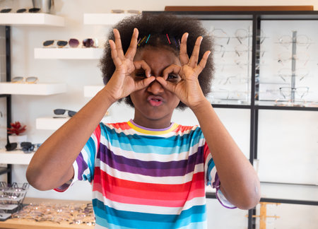 Portrait of a brightly smiling African girl puts her finger in a semicircle symbolizing eyeglasses as she selects a frame in an optical shop.の写真素材